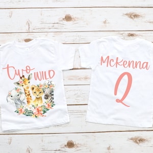 Puede incluir: Dos camisetas blancas con un dise&ntilde;o rosa. La parte delantera de una camiseta dice "Two Wild" con una jirafa, un le&oacute;n, un elefante y una cebra. La parte trasera de la otra camiseta dice "Mckenna 2".