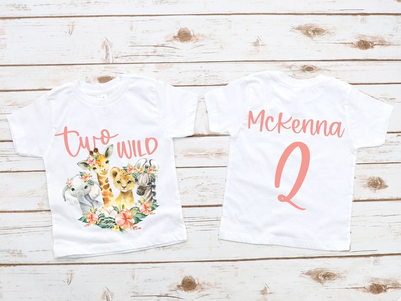Puede incluir: Dos camisetas blancas con un dise&ntilde;o rosa. La parte delantera de una camiseta dice "Two Wild" con una jirafa, un le&oacute;n, un elefante y una cebra. La parte trasera de la otra camiseta dice "Mckenna 2".