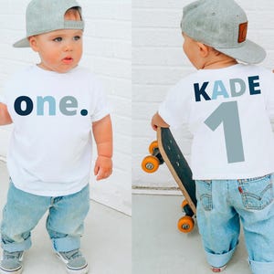 May include: White toddler t-shirt with the word "one" in navy and light blue. The back of the shirt has the name "KADE" and the number "1" in similar colors. The child is wearing light blue jeans and a gray baseball cap.