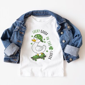 May include: White t-shirt with a cartoon goose wearing a green hat and shoes, riding a skateboard. The shirt has the text "Lucky Goose on the Loose" with shamrocks. A blue denim jacket is draped over the shirt.