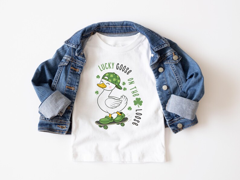 May include: White t-shirt with a cartoon goose wearing a green hat and shoes, riding a skateboard. The shirt has the text "Lucky Goose on the Loose" with shamrocks. A blue denim jacket is draped over the shirt.