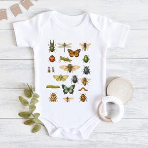 May include: White baby onesie featuring a colorful illustration of various insects, including butterflies, beetles, and ants. The design is printed on a soft, short-sleeved bodysuit, perfect for a baby.