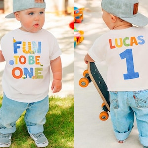 May include: A toddler wearing a white t-shirt with the text "FUN TO BE ONE" in colorful letters. The child is also wearing light blue jeans, gray sneakers, and a gray baseball cap. The back of the shirt has the name "LUCAS" and the number "1".