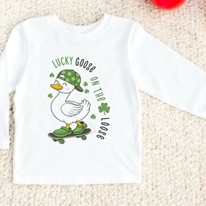 May include: White long-sleeved shirt featuring a cartoon goose wearing a green hat and shoes, riding a skateboard. The shirt has the text "LUCKY GOOSE ON THE LOOSE" in green. The goose has a yellow beak and is smiling.