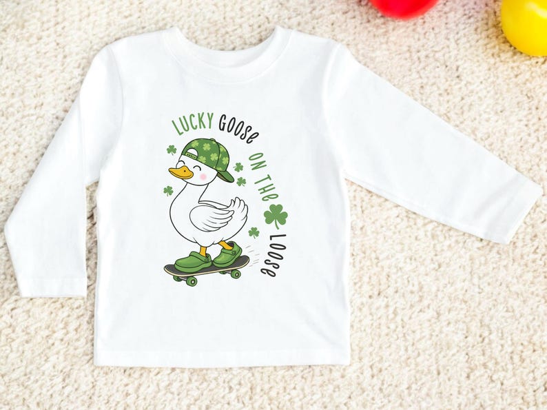 May include: White long-sleeved shirt featuring a cartoon goose wearing a green hat and shoes, riding a skateboard. The shirt has the text "LUCKY GOOSE ON THE LOOSE" in green. The goose has a yellow beak and is smiling.