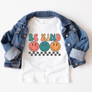 May include: White t-shirt with the text "Be Kind" in colorful letters and three smiling faces. The shirt is layered over a blue denim jacket.