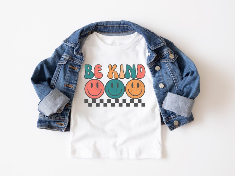 May include: White t-shirt with the text "Be Kind" in colorful letters and three smiling faces. The shirt is layered over a blue denim jacket.