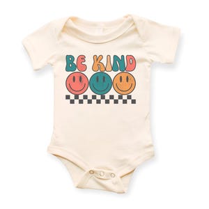 May include: A cream-colored baby onesie with short sleeves features the text "BE KIND" in colorful retro letters with three smiling faces underneath.