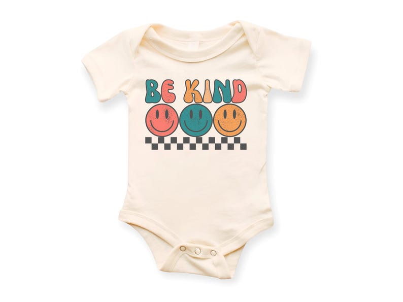 May include: A cream-colored baby onesie with short sleeves features the text "BE KIND" in colorful retro letters with three smiling faces underneath.