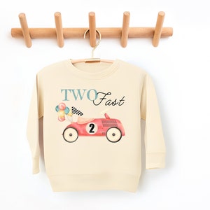 May include: A cream-colored long-sleeved sweatshirt with the text "TWO Fast" and a red toy car with balloons and a checkered flag.