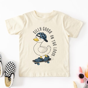 May include: A cream-coloured toddler t-shirt featuring a duck on a skateboard, wearing a blue and black chequered hat and shoes. Text reads "Silly Goose on the Loose". Black and white chequered slip-on shoes are next to the shirt.