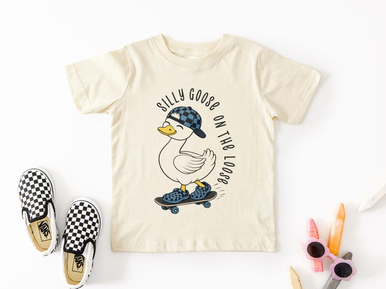 May include: A cream-coloured toddler t-shirt featuring a duck on a skateboard, wearing a blue and black chequered hat and shoes. Text reads "Silly Goose on the Loose". Black and white chequered slip-on shoes are next to the shirt.