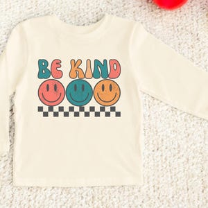 May include: A long-sleeved, cream-colored shirt with the words "Be Kind" in colorful, retro-style lettering. Three smiling faces in different colors are below the text.