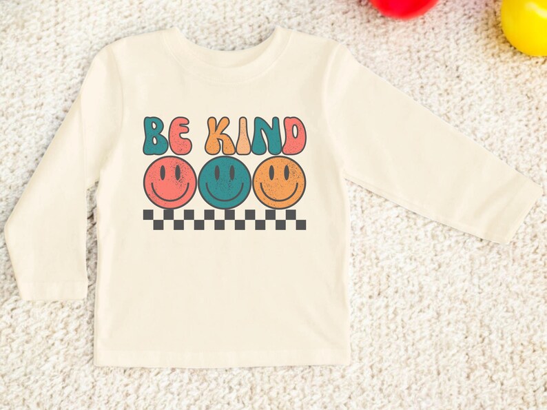 May include: A long-sleeved, cream-colored shirt with the words "Be Kind" in colorful, retro-style lettering. Three smiling faces in different colors are below the text.
