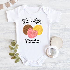 May include: White baby onesie with the text "Tia's Little Concha" in black cursive font. Three colorful concha pastries are pictured below the text.
