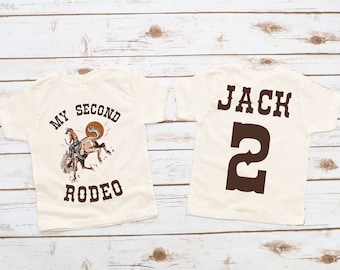 May include: Two white t-shirts with brown text and graphics. The front of one shirt says "My Second Rodeo" with a cowboy riding a bucking horse. The back of the other shirt says "Jack 2".