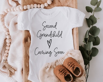 May include: White onesie with the text "Second Grandchild  ♥ Coming Soon" in black script. The onesie is lying in a wooden basket with brown baby shoes and a white blanket.