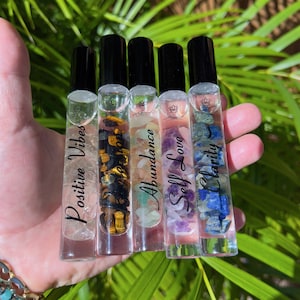 May include: Five clear glass roller bottles with black caps and labels. The labels have different crystal names and affirmations: Positive Vibes, Protection, Abundance, Self Love, and Clarity. Each bottle contains small crystals.