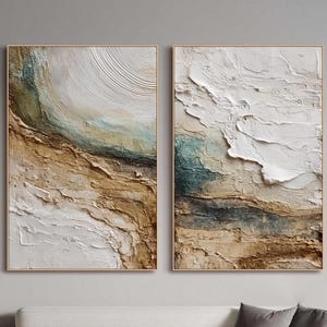 Puede incluir: Dos obras de arte abstractas enmarcadas. Cada una presenta un diseño texturizado y en capas con tonos blancos, marrones y turquesas. La obra de arte se exhibe sobre un sofá blanco con cojines grises, creando una estética de sala de estar moderna.