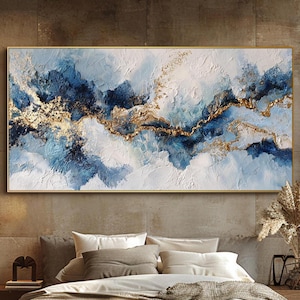 May include: Abstract wall art with a textured painting in shades of blue, white, and gold. The artwork is in a gold frame above a bed with neutral bedding. The room has a modern, minimalist style. The painting is approximately 150cm x 75cm.