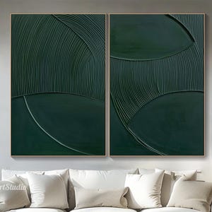 May include: Two abstract paintings in a modern living room. The paintings are a deep green colour and feature a textured, layered design with curved lines. The paintings are hung on a white wall above a white sofa with beige cushions.