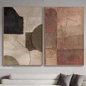 May include: Two framed abstract textured art pieces. One features black, beige, and brown shapes. The other has a reddish-brown and tan colour scheme. Both have a gold frame and are displayed above a white sofa.