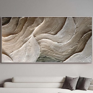 May include: Abstract wall art featuring a textured, layered design in shades of beige, brown, and gray. The artwork is framed and rectangular, with a modern, minimalist aesthetic. The piece is displayed above a white sofa in a living room setting.