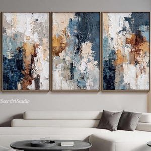 May include: Three framed abstract paintings with textured brushstrokes. The artwork features a palette of white, navy blue, tan, and brown. The paintings are displayed above a white sofa in a modern living room setting.