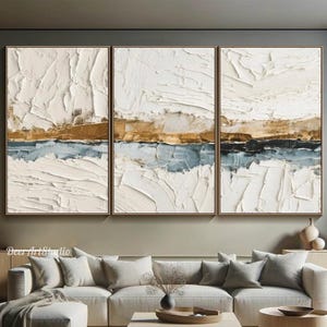 May include: Three framed abstract paintings with textured white, gold, and blue layers. The artwork hangs above a modern living room setup with a sectional sofa, coffee table, and wooden accents. The room has a neutral color palette.