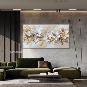 Op de afbeelding: Abstract schilderij met witte en gouden bloemen op een beige achtergrond. Het schilderij hangt aan een muur in een moderne woonkamer met een groene bank en een salontafel.