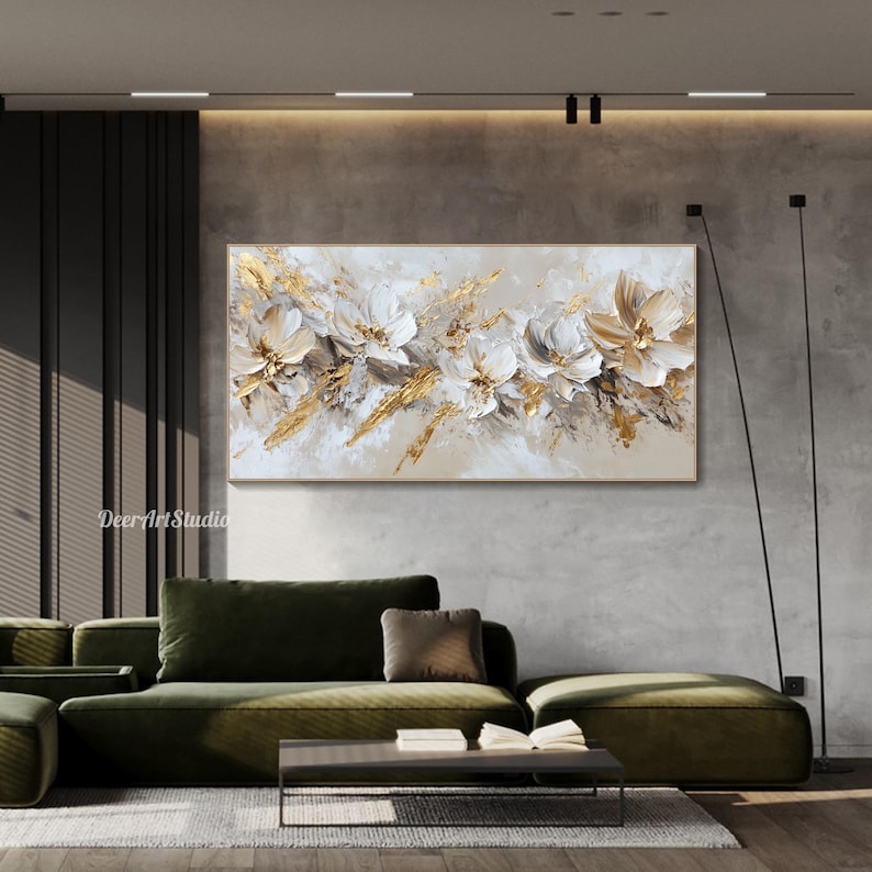 Op de afbeelding: Abstract schilderij met witte en gouden bloemen op een beige achtergrond. Het schilderij hangt aan een muur in een moderne woonkamer met een groene bank en een salontafel.