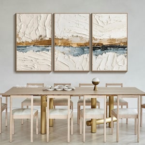 May include: Three framed abstract paintings with textured white, gold, and blue layers. The artwork hangs above a wooden dining table with gold-colored legs and matching chairs. The overall theme is modern home decor.