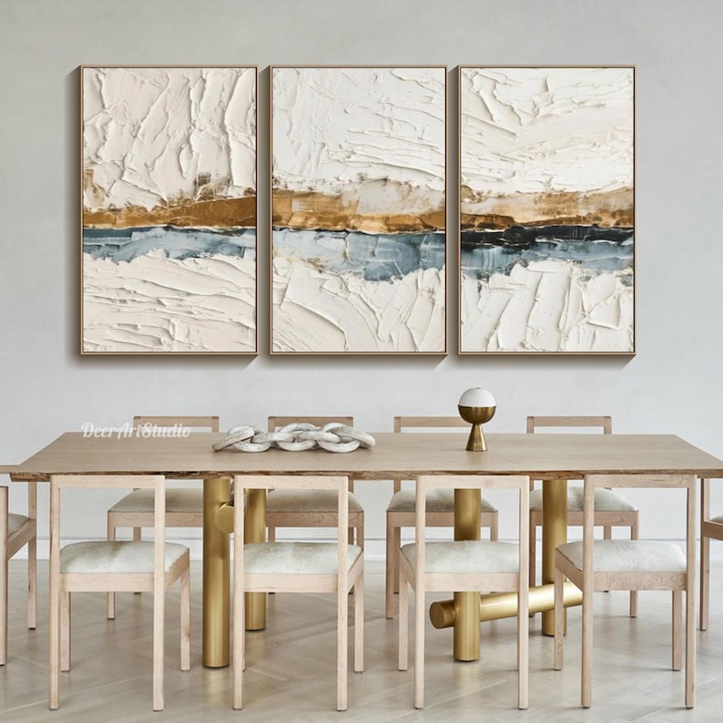 May include: Three framed abstract paintings with textured white, gold, and blue layers. The artwork hangs above a wooden dining table with gold-colored legs and matching chairs. The overall theme is modern home decor.