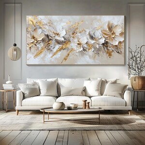 Op de afbeelding: Abstract schilderij met witte bloemen en gouden accenten op een beige achtergrond. Het schilderij hangt boven een witte bank met kussens en een salontafel.