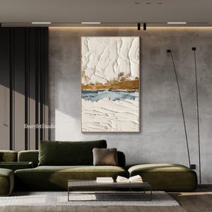 May include: Textured abstract painting with a neutral color palette. The artwork features a layered composition of white, gold, and blue hues, framed in a light wood. The painting is displayed above a green sofa in a modern living room setting.