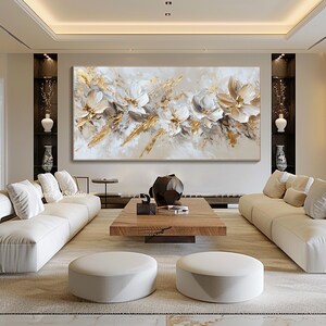Op de afbeelding: Abstract schilderij met witte en gouden bloemen op een beige achtergrond. Het schilderij wordt tentoongesteld in een moderne woonkamer met witte banken en een houten salontafel.