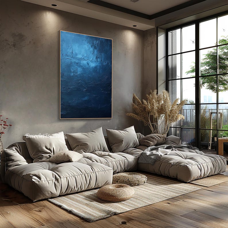 Puede incluir: Una gran pintura abstracta en tonos azules cuelga de una pared en una sala de estar moderna. La pintura est&aacute; sobre un gran sof&aacute; seccional gris claro con una manta y almohadas. La habitaci&oacute;n tiene una ventana grande con vista a los &aacute;rboles y un balc&oacute;n.