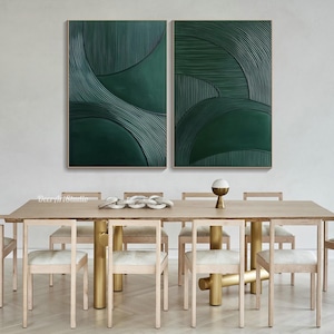 May include: Two framed abstract wall art pieces. Each piece features a deep green textured design with curved and linear elements. The artwork is displayed above a dining table with light wood chairs and a gold-toned base.