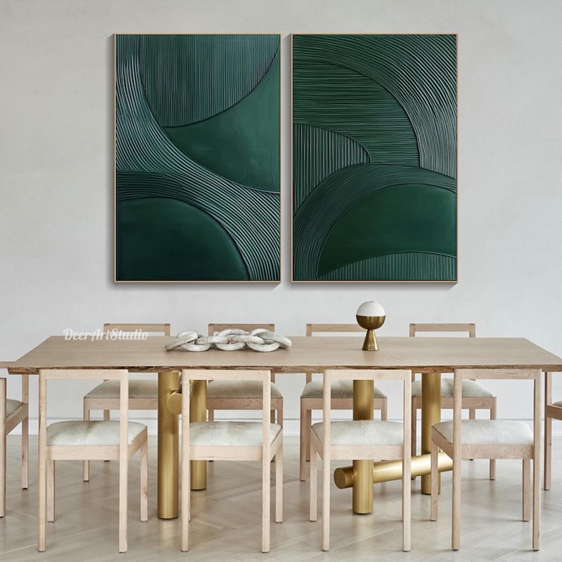 May include: Two framed abstract wall art pieces. Each piece features a deep green textured design with curved and linear elements. The artwork is displayed above a dining table with light wood chairs and a gold-toned base.