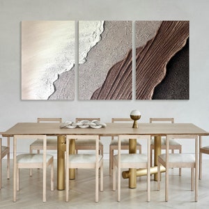 May include: A set of three abstract art prints with a textured, layered look. The prints feature shades of brown, beige, and white, creating a modern and minimalist aesthetic. The prints are displayed above a wooden dining table with gold metal legs.