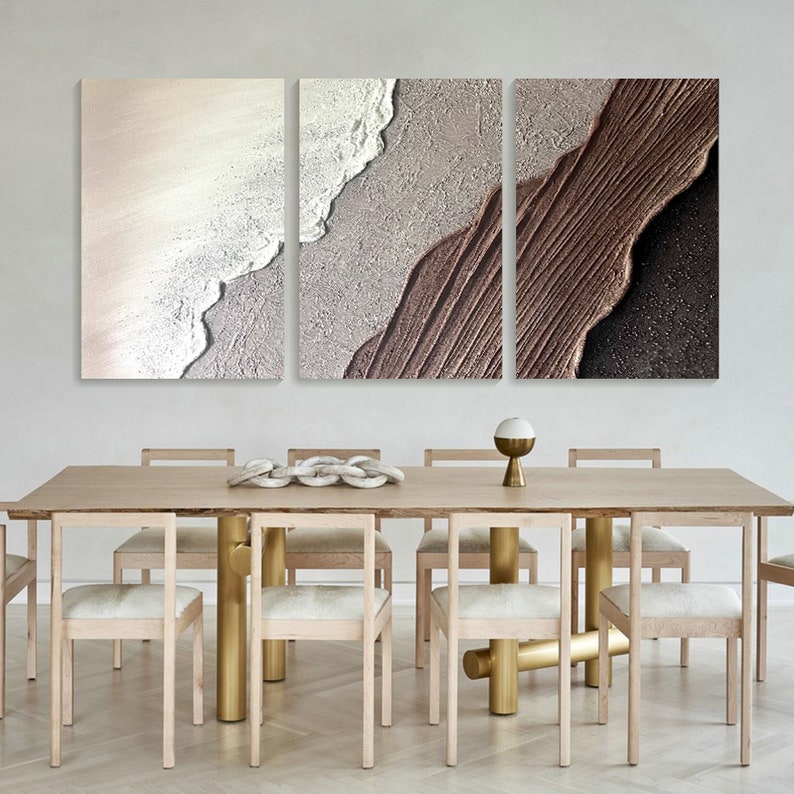 May include: A set of three abstract art prints with a textured, layered look. The prints feature shades of brown, beige, and white, creating a modern and minimalist aesthetic. The prints are displayed above a wooden dining table with gold metal legs.
