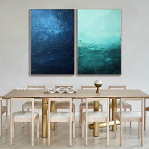 Puede incluir: Dos pinturas abstractas en un comedor moderno. Las pinturas son en tonos de azul y verde, con un efecto texturizado de pinceladas. Las pinturas est&aacute;n colgadas en una pared blanca sobre una mesa de madera con patas doradas. La mesa est&aacute; puesta con seis sillas y un cuenco de objetos decorativos.