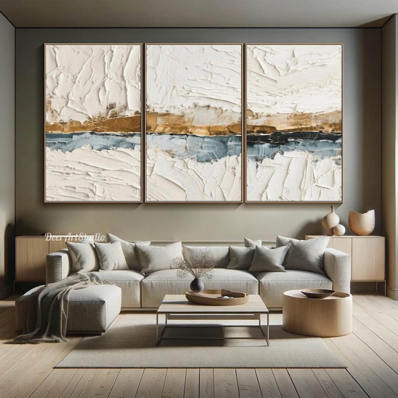 May include: Three framed abstract paintings with textured white, gold, and blue layers. The artwork hangs above a modern living room setup with a sectional sofa, coffee table, and wooden accents. The room has a neutral color palette.