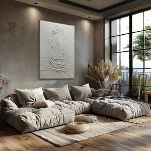 Puede incluir: Un gran sof&aacute; seccional de color gris claro con un dise&ntilde;o acolchado se encuentra en una sala de estar moderna con una ventana grande y una pintura texturizada blanca de una mujer en la pared.