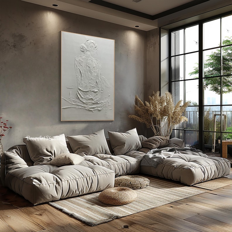 Puede incluir: Un gran sof&aacute; seccional de color gris claro con un dise&ntilde;o acolchado se encuentra en una sala de estar moderna con una ventana grande y una pintura texturizada blanca de una mujer en la pared.