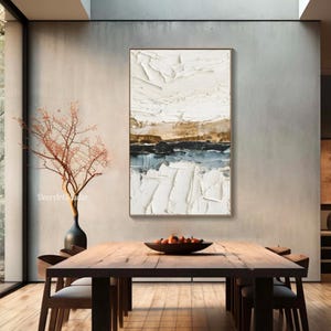 May include: Abstract textured art piece in a gold frame. The artwork features a landscape design with white, brown, black, and blue colors. The painting is displayed above a wooden dining table with chairs.