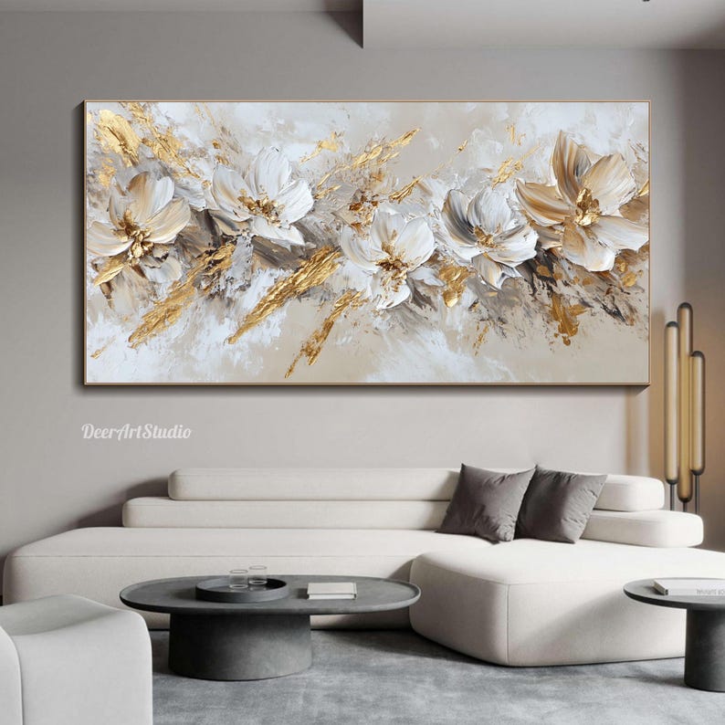Op de afbeelding: Abstract schilderij met witte bloemen met gouden accenten op een beige achtergrond. Het schilderij hangt aan een muur boven een witte bank met grijze kussens.