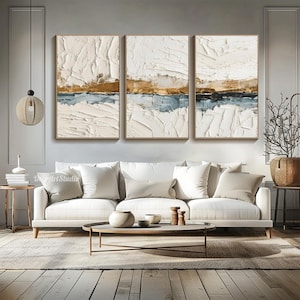 May include: Three framed abstract paintings with textured white, gold, and blue layers. The artwork is displayed above a white sofa in a modern living room setting. The paintings are rectangular and have a neutral color palette.
