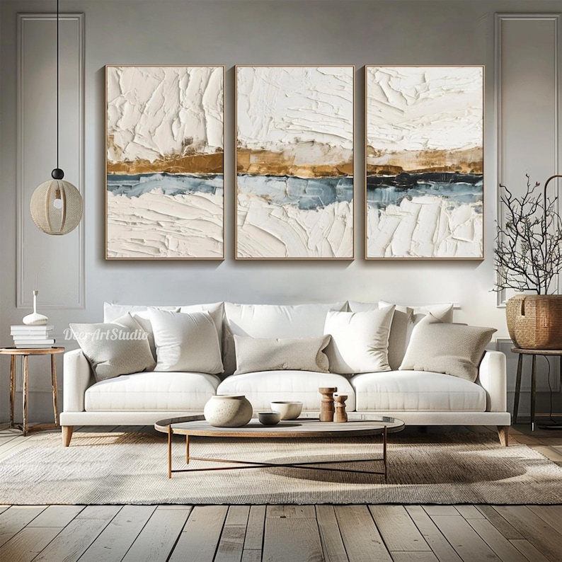 May include: Three framed abstract paintings with textured white, gold, and blue layers. The artwork is displayed above a white sofa in a modern living room setting. The paintings are rectangular and have a neutral color palette.