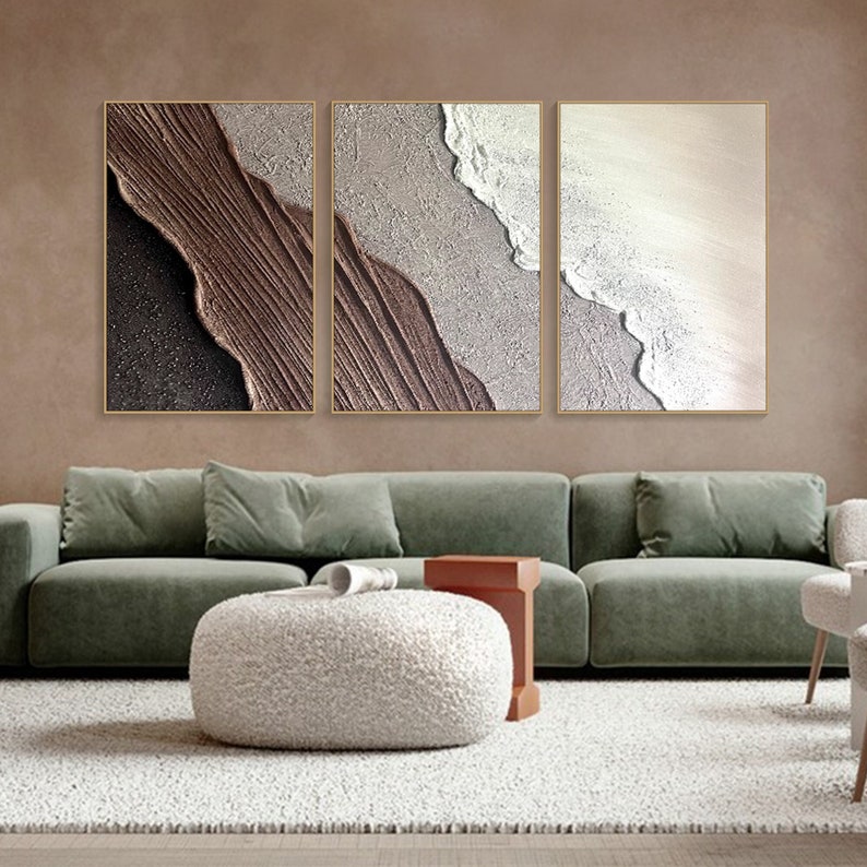 May include: Three abstract paintings in a living room setting. The paintings are in a modern style and feature a brown, white, and beige color scheme. The paintings are hung on a wall above a green sofa and a white coffee table.